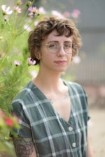 Margot fisher is an author with short curly brown hair, round glasses, and green dress standing in front of a flowering bush