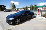 A black Model S Tesla sedan charges at a Tesla supercharging station on a sunny day.