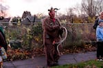 A man dressed in a furry suit and with a red face mask with a long tongue stands on the sidewalk.