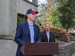 Oregon Senator Ron Wyden, wearing a blue suit and baseball cap, speaks from a podium.