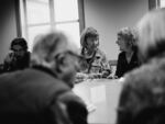 In this black-and-white photo, Kim Butler is on the left and Pam Blakeman is on the right. Both are seated next to one another at a conference table and are looking at one another as they speak together. Other people are also seated around the table.