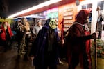 People dressed in creepy costumes and masks walk in a procession. The person in front is wearing a red hood and carrying a staff with a bell, and the next person in line is wearing a green monstrous mask with a wide, toothy smile.