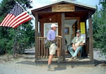 a wilderness ranger with a white beard and green hat sits on the porch of a small cabin and talks to a hiker wearing a purple shirt and fanny pack