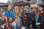 Ten band members stand outside posing for a picture. Everyone is smiling and holding an instrument, like different types of horns and drums. In front, a band member wrapped in a Pride flag holds a rainbow megaphone.