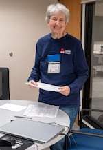 A volunteer with grey hair and a smile stands holding papers and wearing a long-sleeved blue tshirt in an office. A laptop is visible in the front.