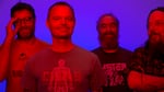 Four people stand together with slight smiles on their faces. The lighting is distorted so they appear to be very reddish in color, and the background is a bright purplish blue.