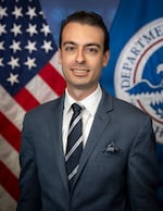 This photo shows Paul Ingrassia photographed from about the waist up. He's wearing a suit and tie, and behind him are an American flag and a Department of Homeland Security flag.