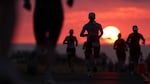 With a red sunset in the background, triathletes photographed in silhouette run in the final portion of the 2025 Ironman World Championship.