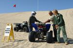 An ATV rider wearing a helmet stands to the left of an ATV while two people wearing U.S. Forest Service uniforms inspect it. They are on sand next to a sign that reads, "SOUND MONITORING STATION."