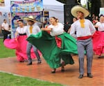 A group of young dancers performing on stage. The girls are wearing white blouses and big colorful skirts that they're swinging as they dance, with flowers in their hair. The boys are wearing sombreros, white embroidered shirts with yellow ties, striped pants and boots. Food vendors are visible past the stage.