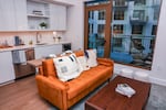 An orange couch and coffee table are set up near a small kitchen with a view of glass doors that open to a small patio overlooking another high-end residential building.