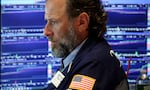 A trader works on the floor of a stock exchange. He is wearing a navy blue blazer, and screens displaying stock market information are behind him.