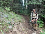 A man stands on a trail in a forest. He has a backpacking pack on and a compound archery bow.
