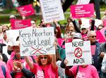 People wearing pink hold up signs supporting abortion rights. Signs include images of wire hangars crossed out and words like "Keep abortion safe and legal"