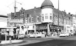 A black-and-white photo shows a bustling business district with a lunch counter, drug store, cafe, dry cleaner and other community services.