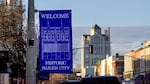 A blue sign on a lightpost states "Welcome Historic Baker City". The historic Geiser Grand Hotel and Baker Tower loom in the background.