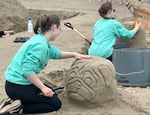 A girl with her back turned smiles and sculpts a pumpkin with the face of a pug. Another girl behind her does the same. Both are wearing matching team sweatshirts.