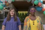 Two women in brightly colored clothes stand side-by-side in front of a house and balloons.