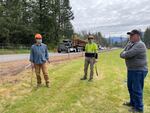 Three men are standing next to a tree-lined highway. Two are holding axes.