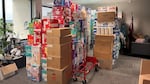 Pictured is the corner of an office, filled with boxes of diapers, toilet paper and other hygienic goods. Brands that can be see are Huggies in it's red box, Pampers in it's aqua blue and several unlabeled brown boxes. The piles of the boxes reach the ceiling and take up a good portion of the room.