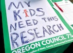 A handwritten sign rests on a table at a rally supporting higher education researchers in Portland on Feb. 19, 2025. Researchers at Oregon universities say efforts to defund their work will have negative impacts on local communities.