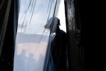 A transgender man living wearing a baseball cap is shown in silhouette in a doorway behind a sheer curtain.