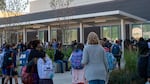 A dozen or more adults and students can be seen from behind as they face a school building and walk towards it. Many parents hold their chlidren's hands or hug them, and many children wear backpacks.