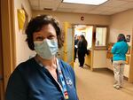 A person wearing medical scrubs with the words "Coquille Indian Tribe" on them wears a mask and looks a the camera, while people stand in line behind her, maintaining a distance of 6 feet between each group.