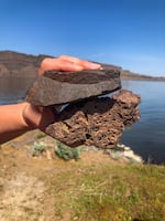A hand is holding a black rock with holes in it underneath another black rock that is solid. Behind the rocks, you can see brown and green grass at the bottom of the picture, a blue river in the middle of the picture, brown rocky hills across from the river, and a blue sky.