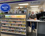 A pharmacist fills out paperwork behind a counter that's next to a shelf full of vitamins and medicines.