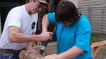 Shop teacher Chris Mathas helps Kaden Delatorre drill holes in an oak log.