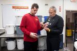 A young man in a red shirt puts on gloves, with help from a man with graying hair in a blue shirt. They stand in front of white tubs in the popcorn store.