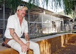A man with tattooed arms and a baseball cap on his head sits on a low outdoor wall with animal cages visible in the background.
