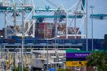 Very large industrial cranes loom over stacked containers with an enormous ship filled with containers visible in the background.