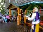 A man in Nez Perce regalia plays a handheld drum and sings into a microphone on the wooden deck of an old hotel as an audience quietly listens.