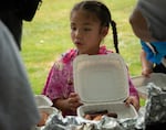 A young girl with braids has a takeout tray with slices of eel-like fish at a buffet table.