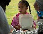 A young girl with braids has a takeout tray with slices of eel-like fish at a buffet table.