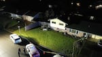 A photo taken from above shows police tape cordoning off a yard and house. Inside the tape, tire tracks show the path a car took before it crashed into the fence of the house. Outside the tape, three police vehicles are parked, and three officers stand in the street and talk to one another.