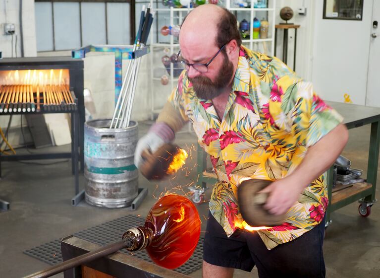 A man in a floral shirt holds flaming oven mitts to either side of a round piece of hot glass.