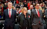 Three people, two men with a woman between them, stand courtside while watching a Portland Trail Blazers basketball game.