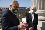 Chuck Schumer, wearing eyeglasses, a face mask and a suit, holds a paper in his left hand and gestures with his right hand while he speaks to someone off camera while standing outside of the U.S. Capitol. Karen Gibson crosses her hands in front of her as she listens.