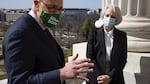 Chuck Schumer, wearing eyeglasses, a face mask and a suit, holds a paper in his left hand and gestures with his right hand while he speaks to someone off camera while standing outside of the U.S. Capitol. Karen Gibson crosses her hands in front of her as she listens.