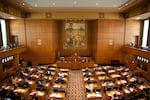 The floor of the Oregon of Representatives, from above and behind state legislators, shows people seated at desks facing the front of a room, while House leaders face the rest of the people assembled. In high balconies, other people sit to observe proceedings.