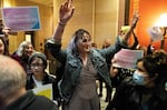 Rep. Leigh Finke, Minnesota's first openly trans legislator, has curly medium-length hair dyed light purple. She is wearing a jean jacket and has her arms raised in celebration. Young people around her carry signs that say, "Pass the trans refuge bill."