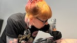 A woman with orange, red and purple dyed hair, leans over tattooing on a person's leg. Her arms are both covered in tattoos.
