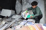 In this photo, a boy wearing black pants and a dark green top looks through the rubble of a building and is lifting out books.