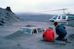 Two man are seen from behind examining a car buried up its windshield in gray ash. A helicopter and a rocky ridge are visible in the near distance.