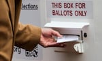 A hand puts a ballot envelope into a slot in a box with text that reads "This box for ballots only."