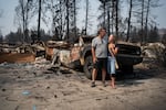Ken and Shannon Adkins stand in front of their home, which was destroyed by wildfire. A heavily damaged truck sits behind them.