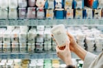 A close shot of a woman's hands holding a carton of yogurt while standing in the dairy section of a supermarket.